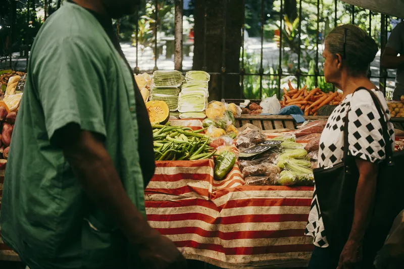 Barraca de feira com legumes e frutas; um homem e uma senhora passam pela banca.