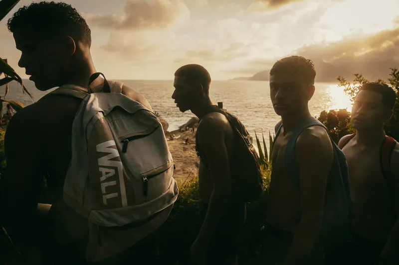 Garotos com mochilas enfileirados contra o sol baixo, à beira do mar, no Rio de Janeiro.
