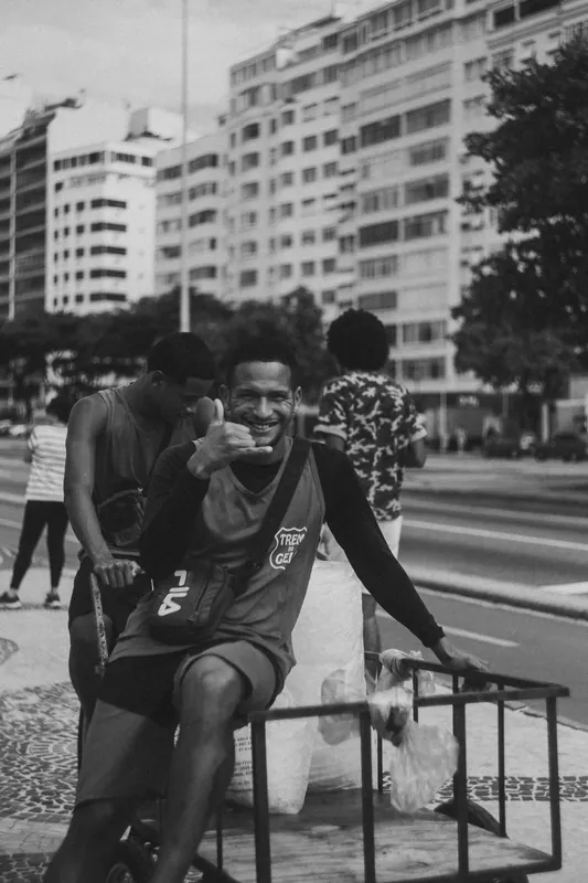 Vendedor sorrindo e fazendo sinal com a mão em frente aos prédios de Copacabana, em preto e branco.