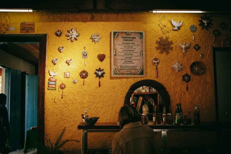 Parede amarela de um bar coberta de divinos e um quadro com uma oração.