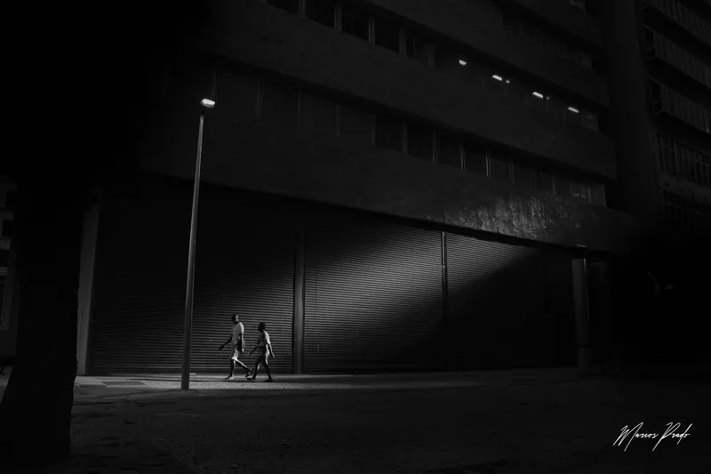 Duas pessoas atravessando um facho de luz de poste à noite, em preto e branco.