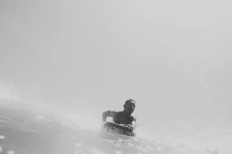 Rapaz remando na prancha de bodyboard dentro do mar, quase todo branco, em preto e branco.
