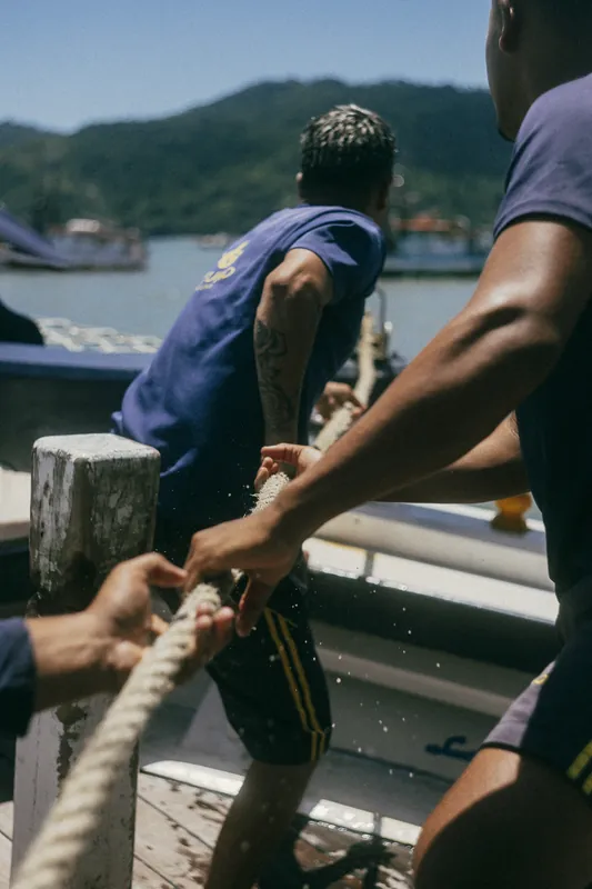 Mãos puxando um cabo de amarração no convés de uma embarcação.
