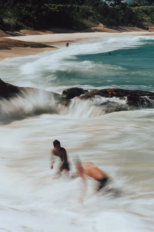 Garotos brincando na espuma da onda em uma praia de pedras, com a água em movimento arrastado.