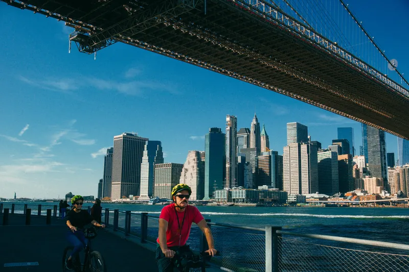 Ciclistas passando sob a ponte do Brooklyn com o skyline de Manhattan ao fundo.