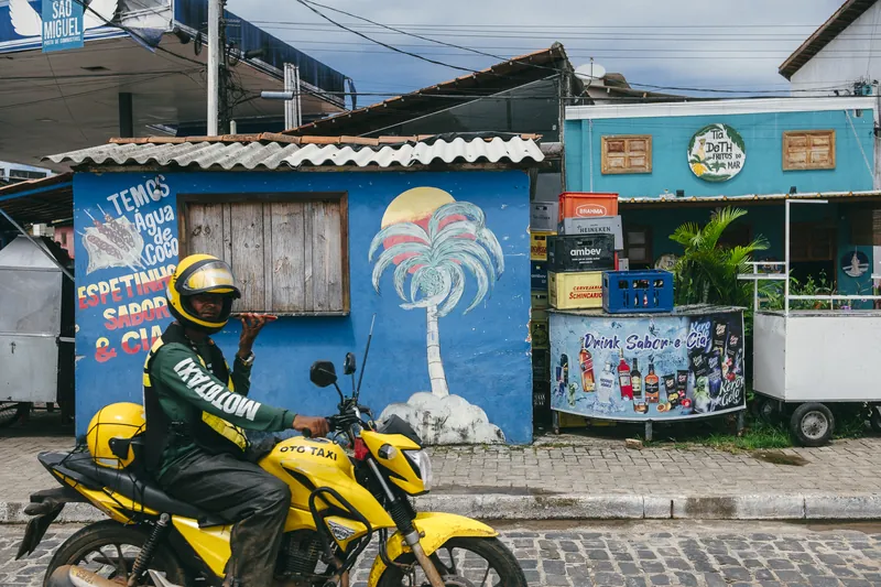 Mototaxista parado diante de um muro azul pintado com coqueiro.
