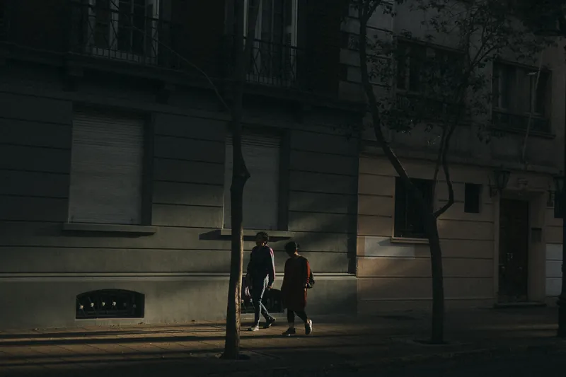 Duas mulheres caminhando por uma calçada iluminada pelo último sol, entre prédios escuros.