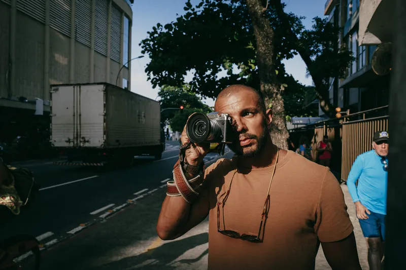Marcos Prado fotografando na rua com uma Fujifilm X-T5 junto ao rosto, no fim de tarde.