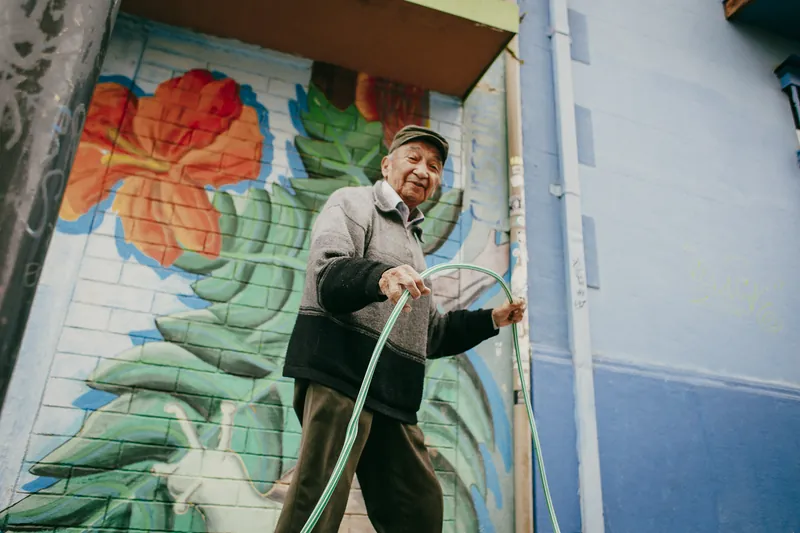 Senhor de boina enrolando uma mangueira na calçada, diante de um muro pintado com flores.