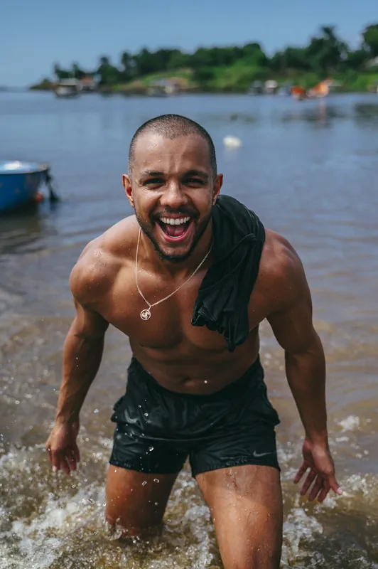 Marcos Prado saindo da água, rindo, com a camiseta no ombro e barcos ao fundo.