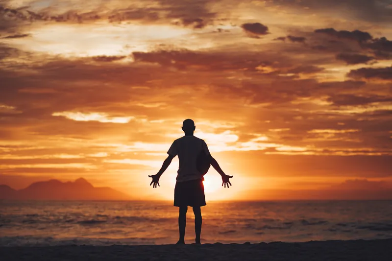 Homem de braços abertos diante do mar, contra o sol nascendo entre as nuvens.